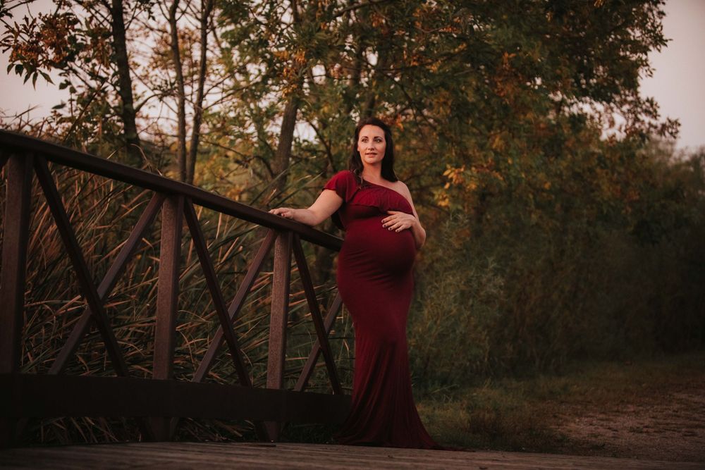 Expectant mother in elegant red gown poses on rustic bridge during sunset.