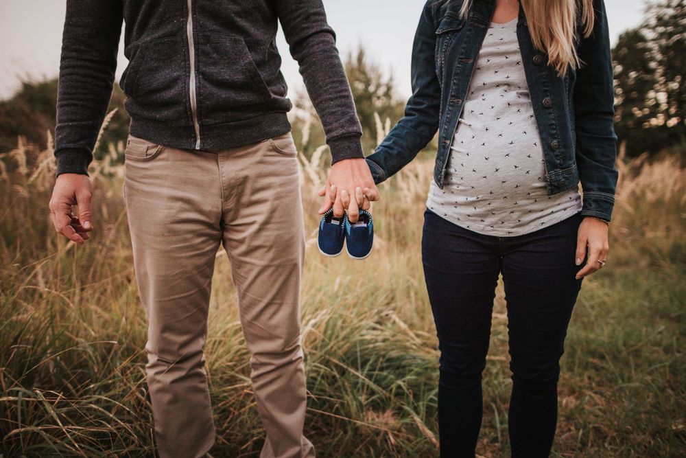 Expectant couple holding tiny blue baby shoes while standing in a grassy field at sunset.