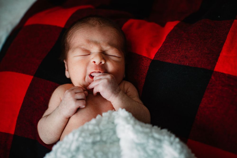 Sleeping newborn baby on red buffalo plaid blanket with hand near mouth.