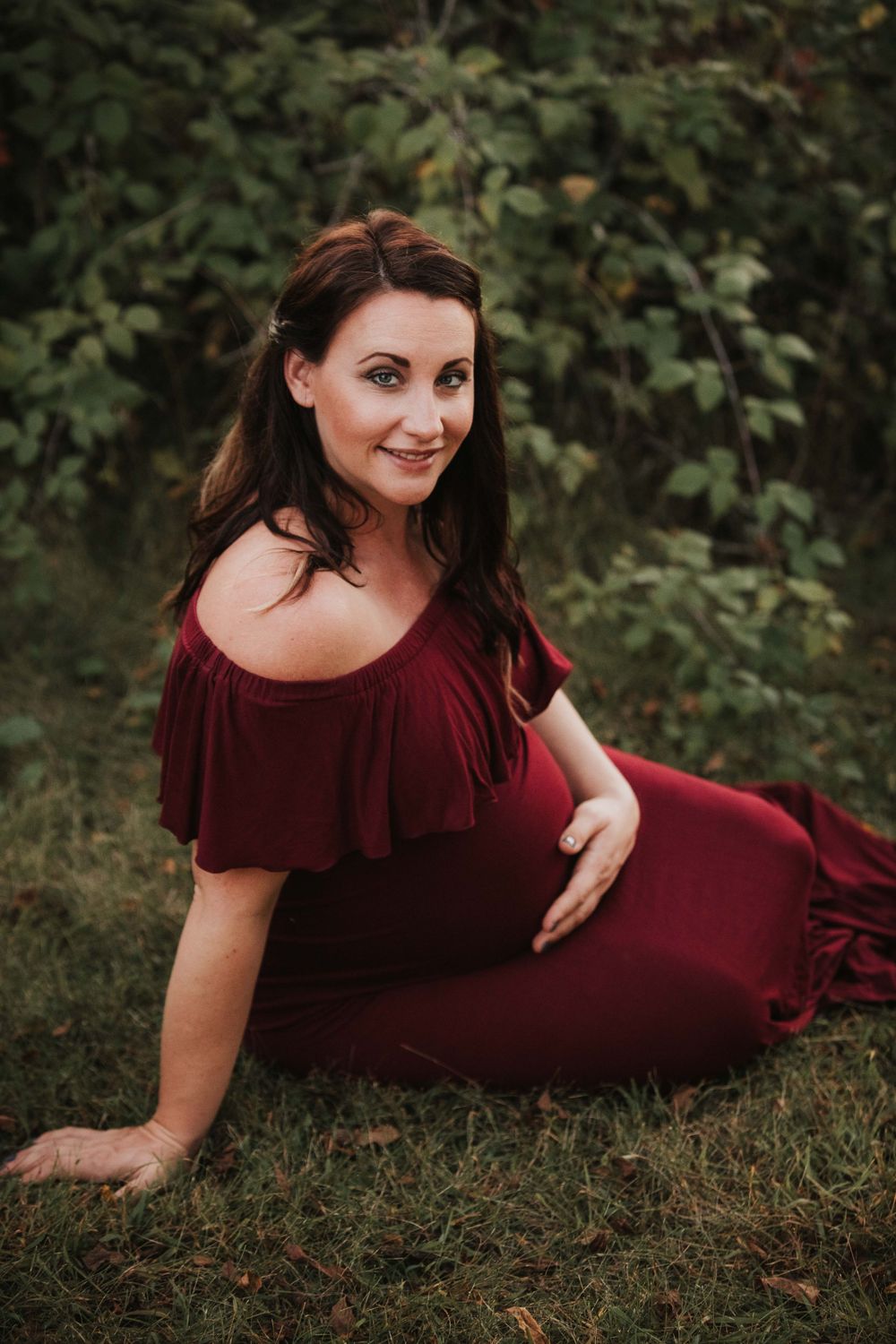Maternity photo of woman in burgundy dress sitting on grass during outdoor photo session.