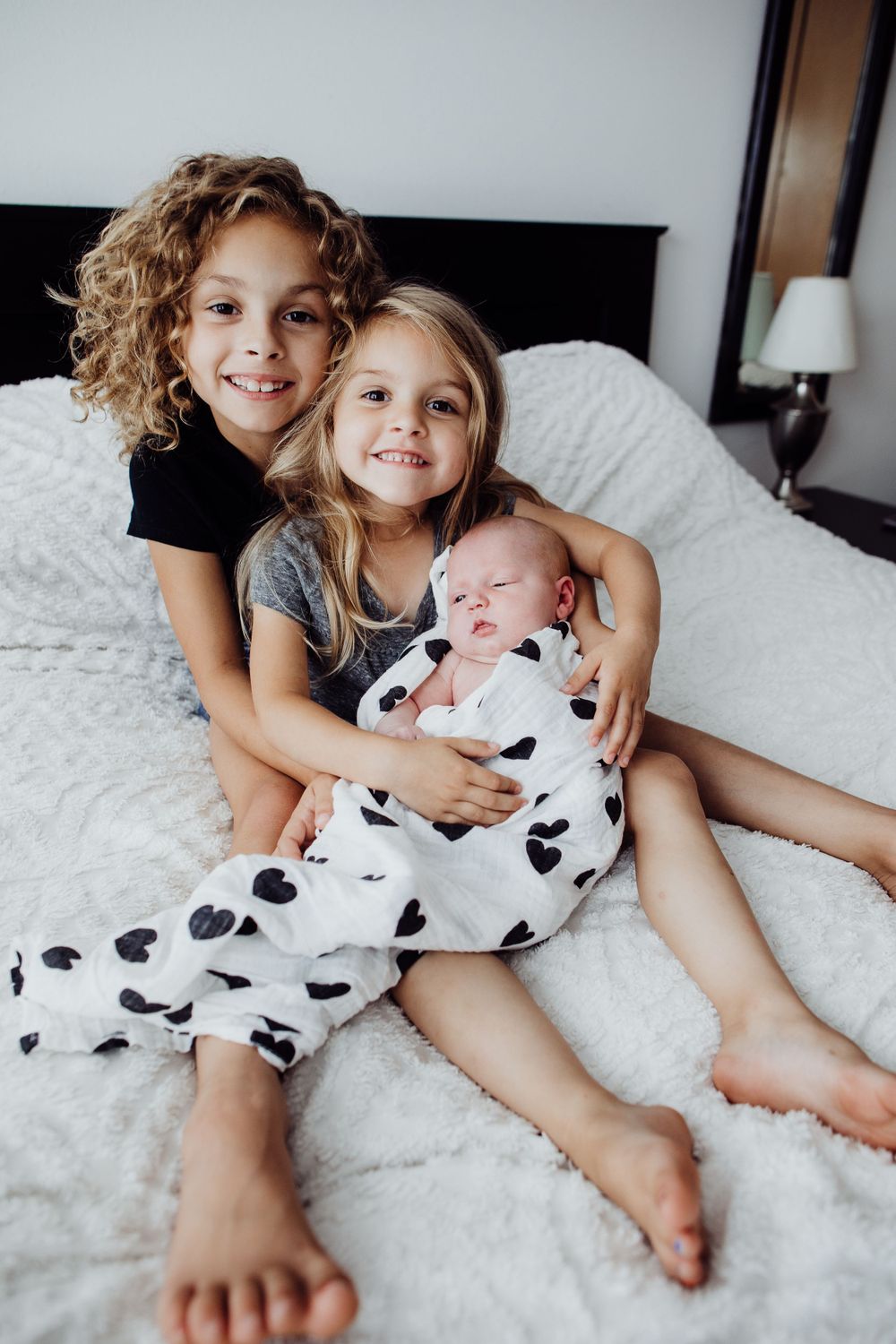 Three siblings cuddling together on white bed with newborn in cow print outfit.