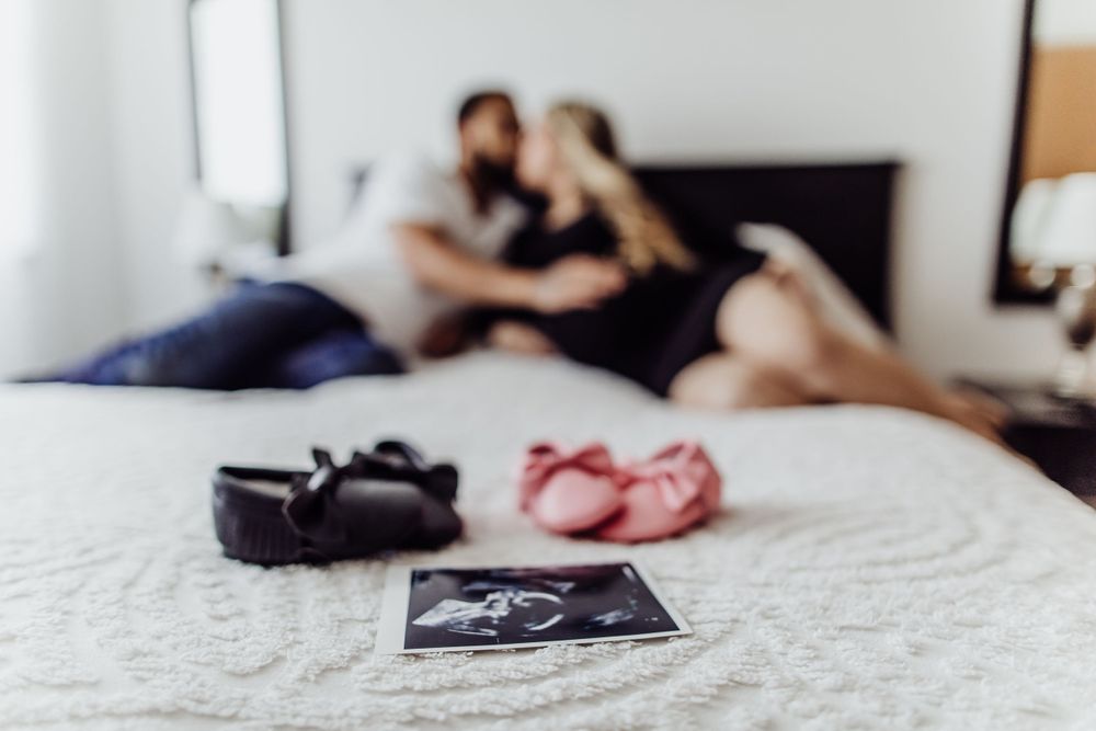 Maternity photoshoot on bed with ultrasound photo and baby shoes in foreground.