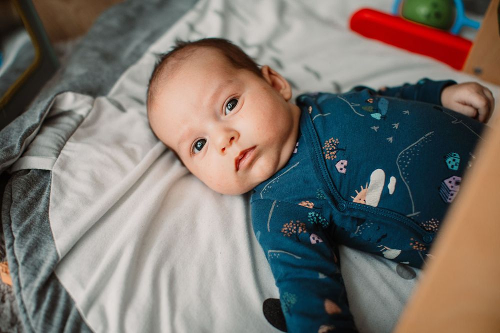 Baby wearing navy blue patterned onesie lying on white bedding.