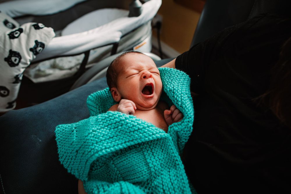 Newborn baby wrapped in turquoise knit blanket crying with eyes closed.