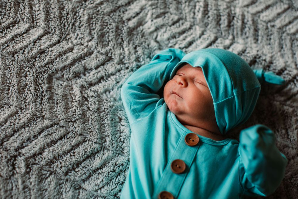 Newborn baby wearing turquoise outfit lying on textured gray blanket.