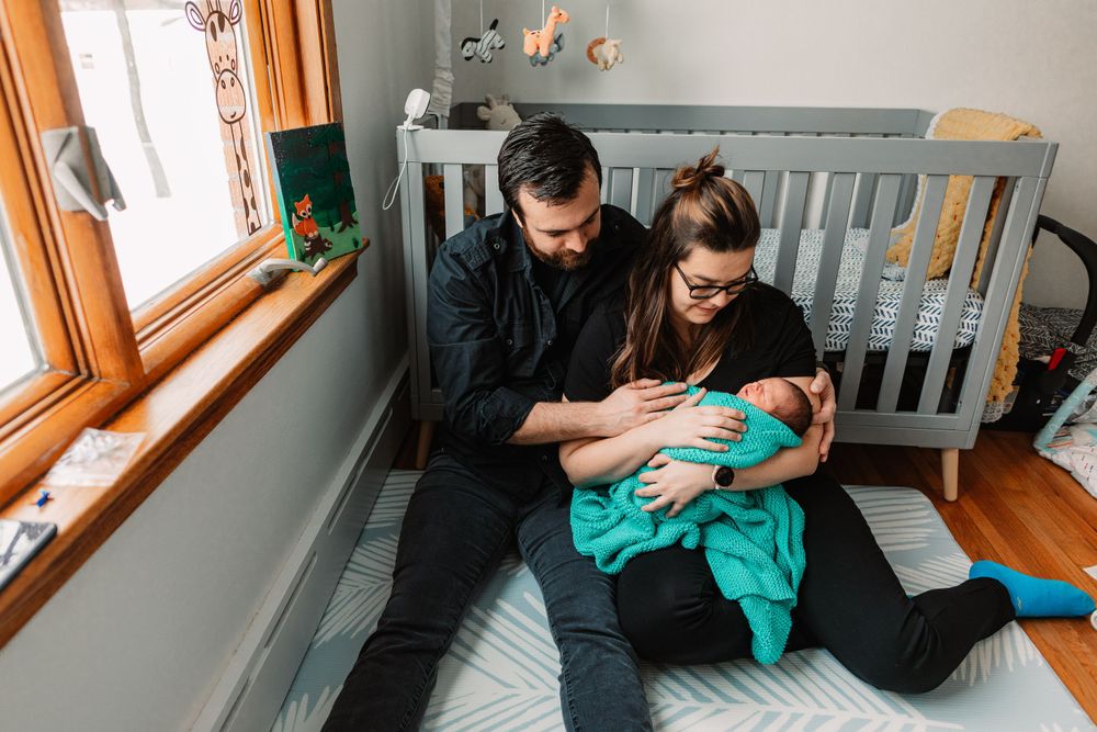 Family moment in nursery with gray crib as couple holds newborn in teal blanket.