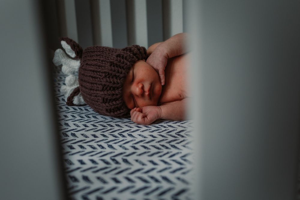 Sleeping newborn wearing brown knitted hat and outfit on patterned bedding.