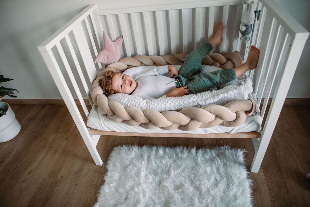 Toddler lying in white crib with braided cushion and plush toys.