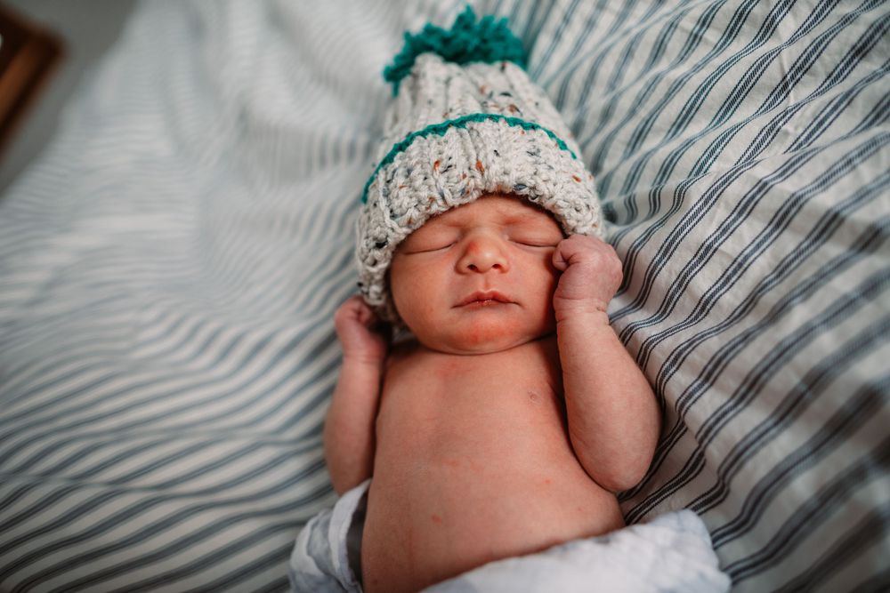 Newborn baby wearing mint green knit hat stretches while lying on striped bedding.