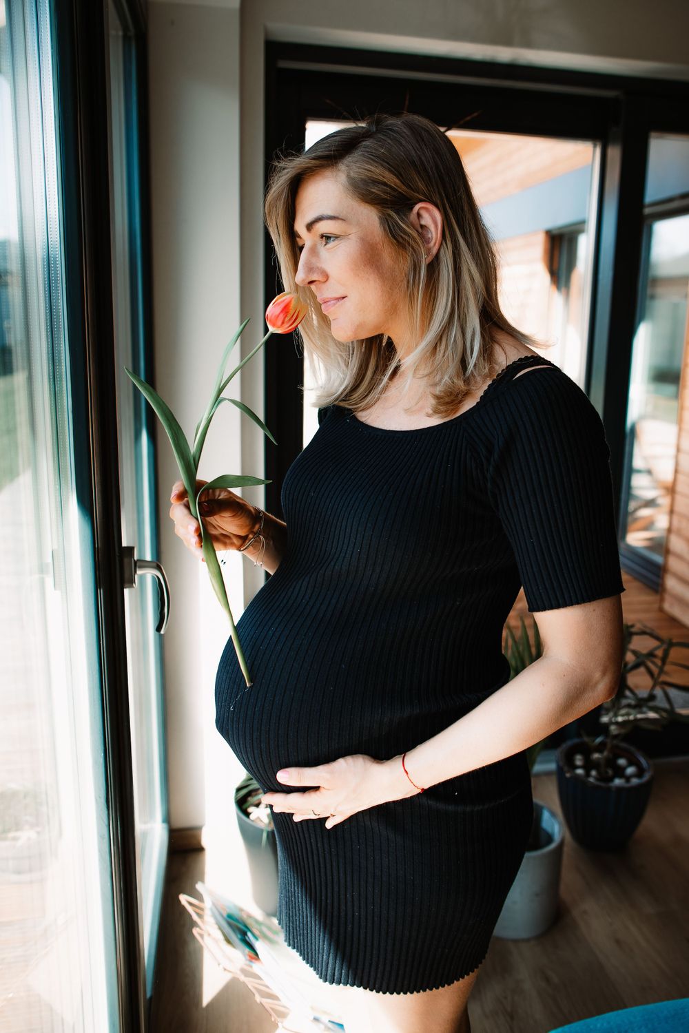 Expectant woman in black dress holding flower by window in sunlit room.