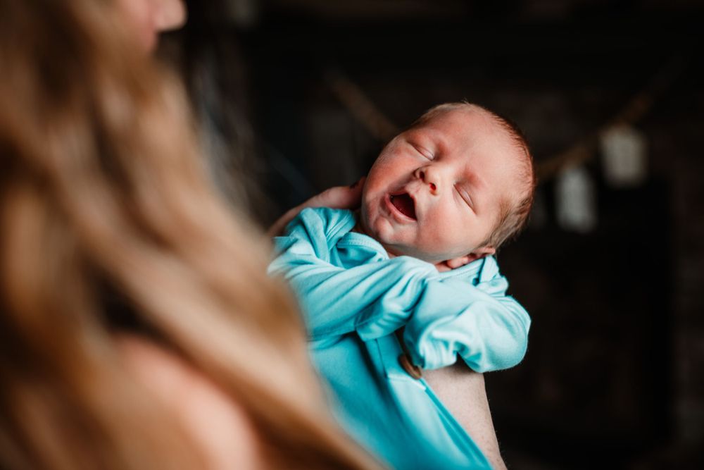 Newborn baby wearing light blue sleepwear cries while being held against dark background.