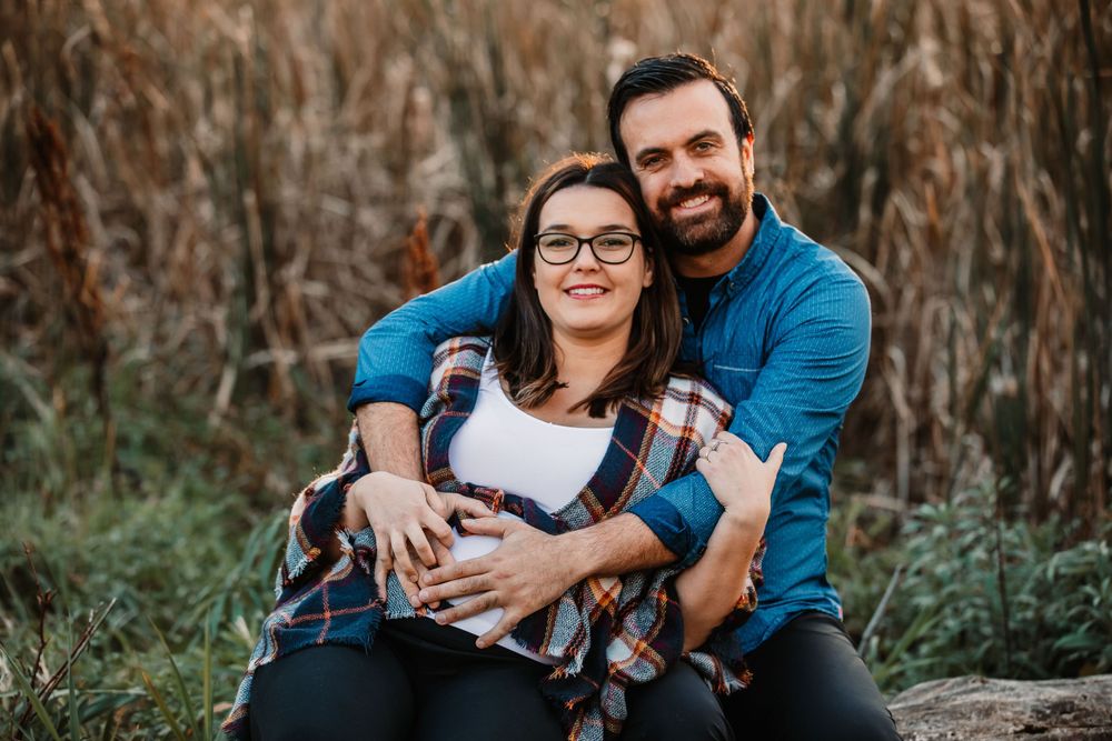 Happy expectant couple in casual attire sitting together outdoors against autumn backdrop.