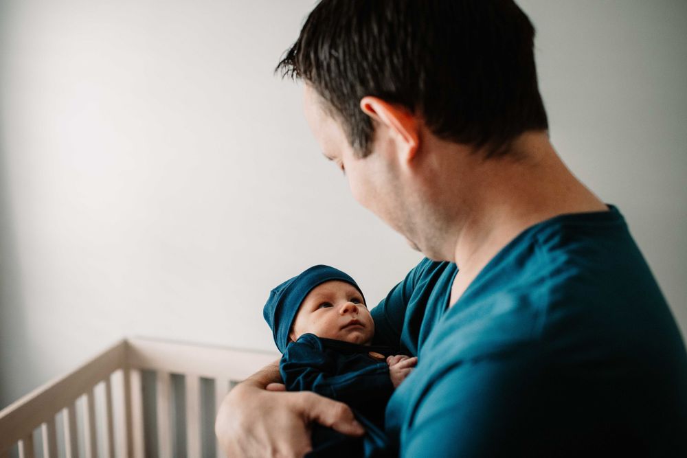 Person wearing blue shirt holds newborn baby near white crib.