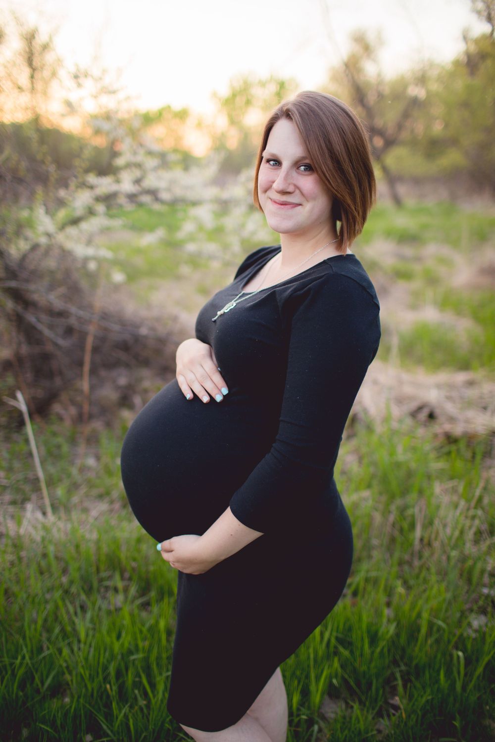 Expectant mother in black dress poses in natural outdoor setting.