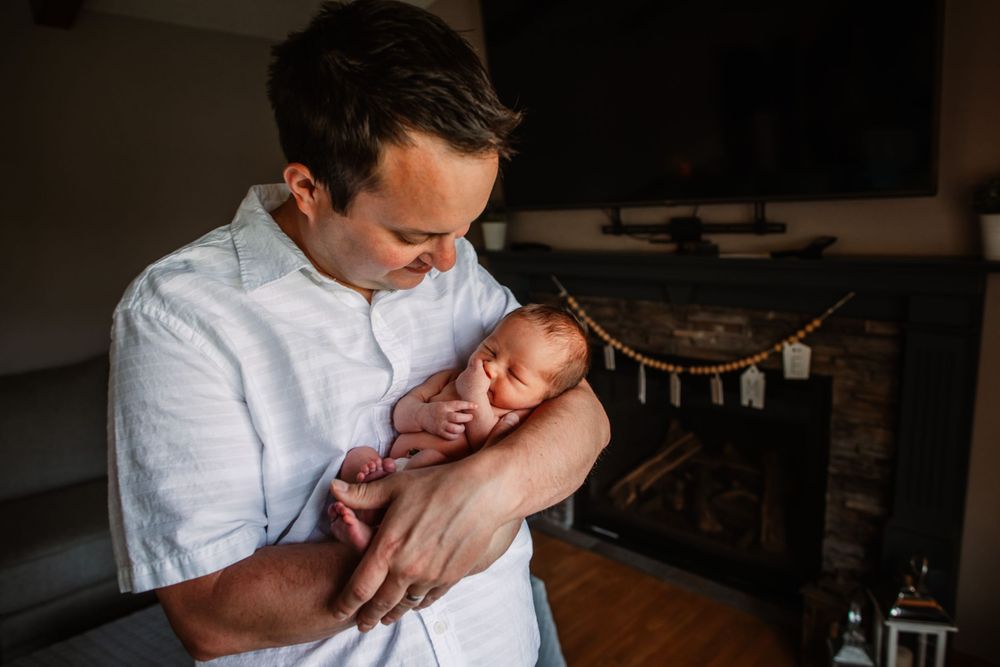 Parent cradling sleeping newborn baby while sitting indoors near fireplace.