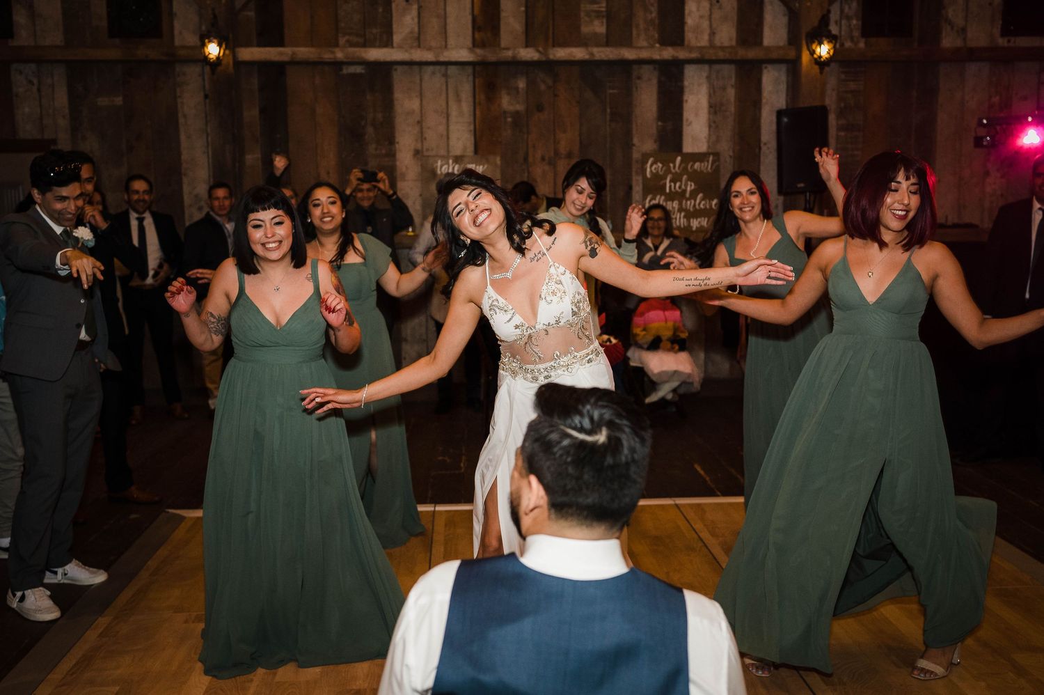 Wedding party members dance in sage green dresses on a wooden dance floor.