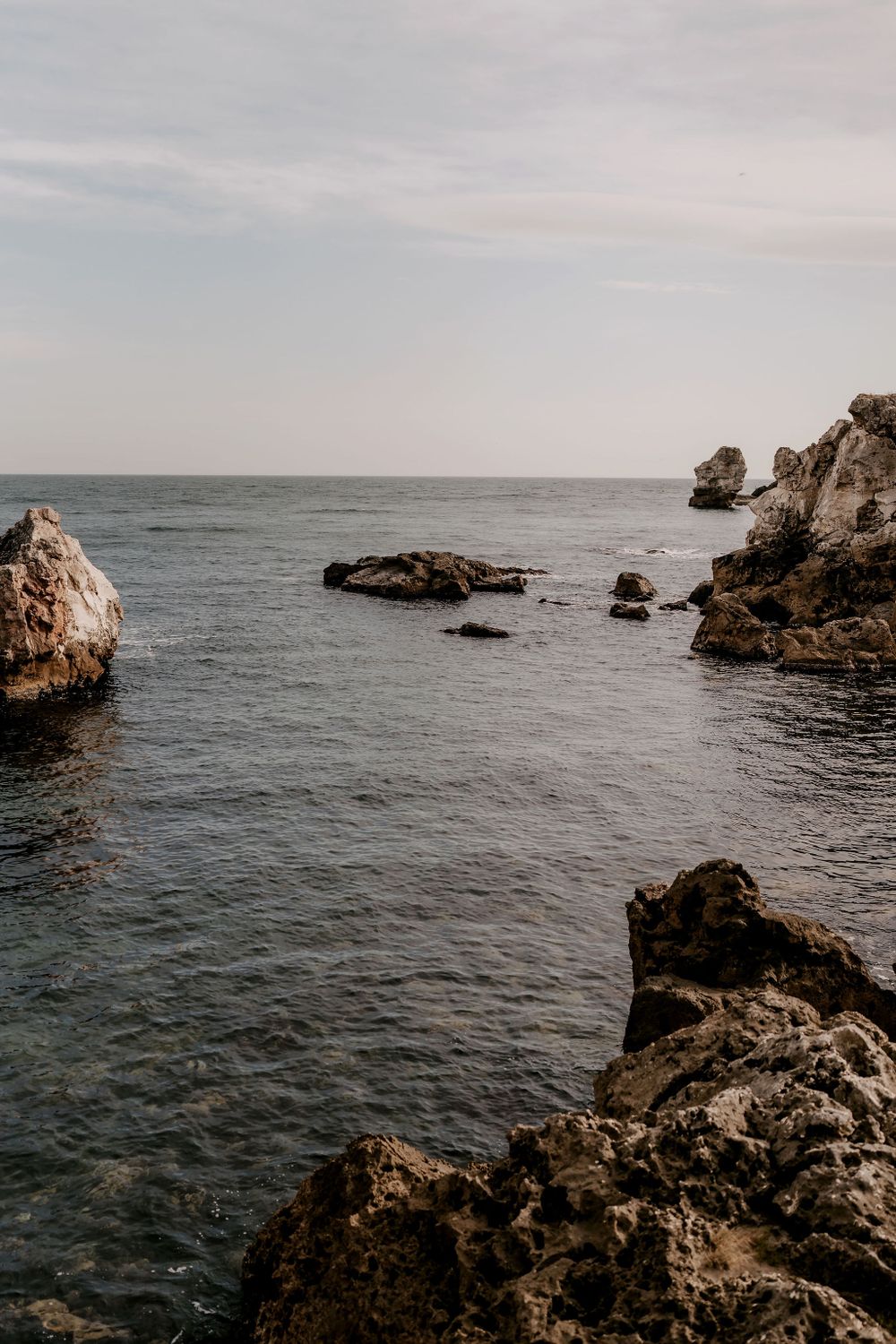 Rocky coastline meets the ocean under an overcast sky with waves crashing against jagged cliffs.