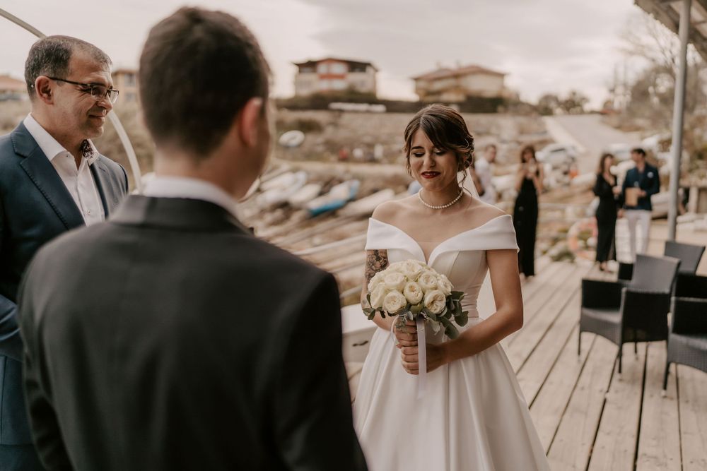 A wedding ceremony takes place on a scenic waterfront deck with guests seated in the background.