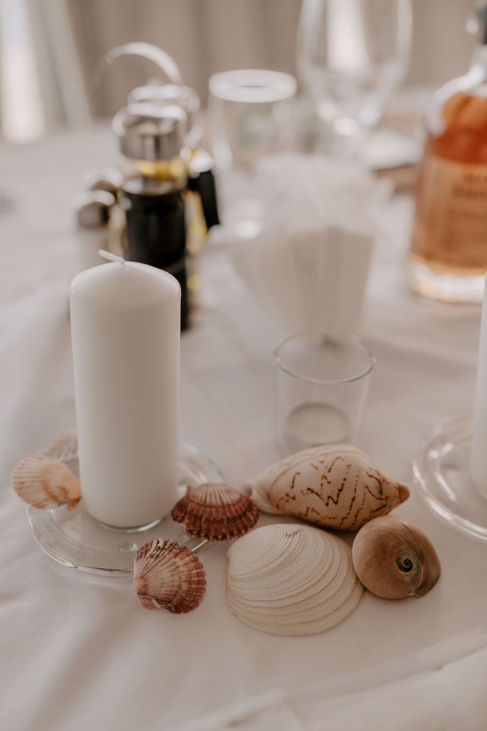 Coastal-themed table decor with white candles and seashells on crisp white tablecloth.