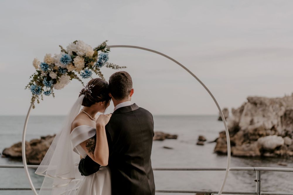 Wedding couple shares a moment under floral arch with coastal rocks and water in background.