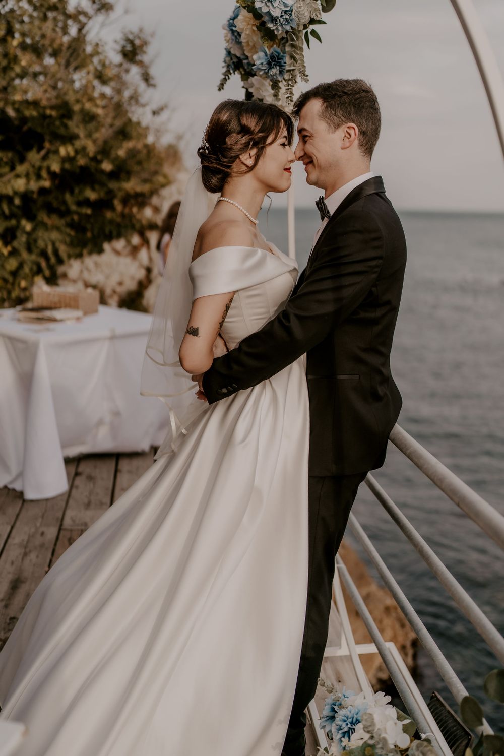 A romantic wedding couple embraces on a waterfront deck with an ocean view at sunset.