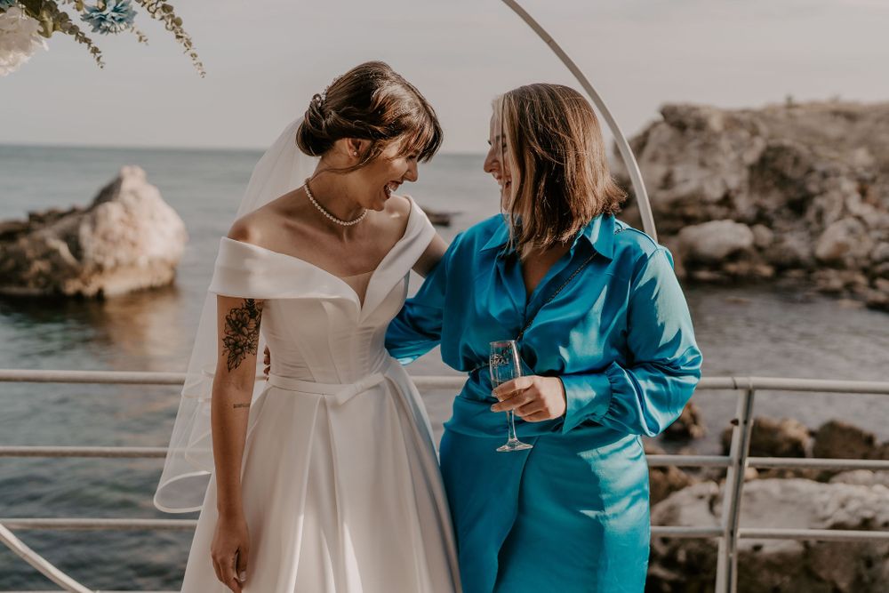 A bride in a white off-shoulder gown stands with a guest in blue on a seaside deck.