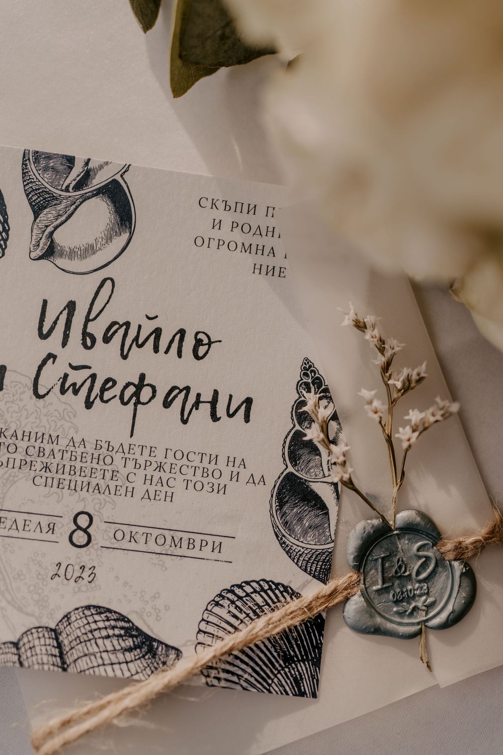 A wedding invitation with delicate illustrations and wax seal lies among dried flowers.
