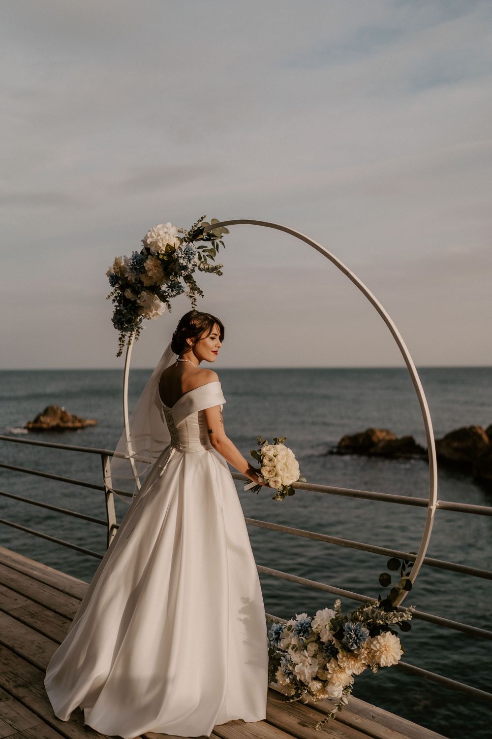 A bride in an elegant off-shoulder gown poses under a floral arch against an ocean backdrop.