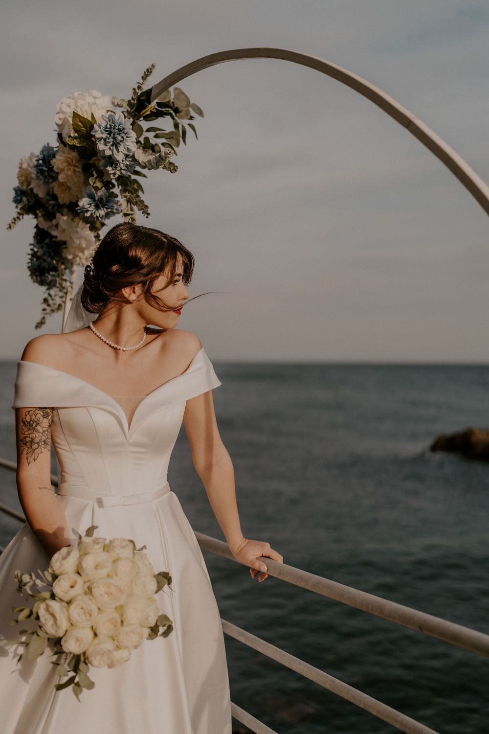 A bride in an off-shoulder gown holds a white bouquet under an arched floral installation.