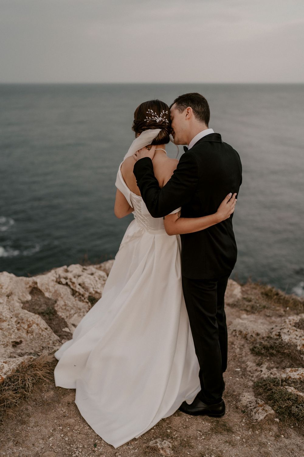 A romantic wedding couple embraces on a rocky cliff overlooking a moody ocean at sunset.