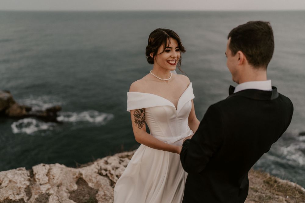 Elegant couple in wedding attire share a moment on rocky coastal cliffs.