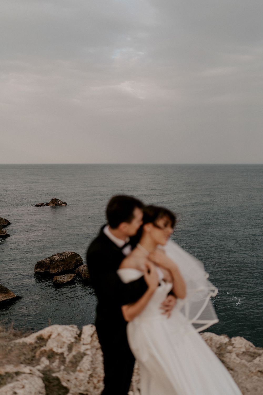 Dreamy wedding portrait with blurred couple against rocky coastline and misty sea.