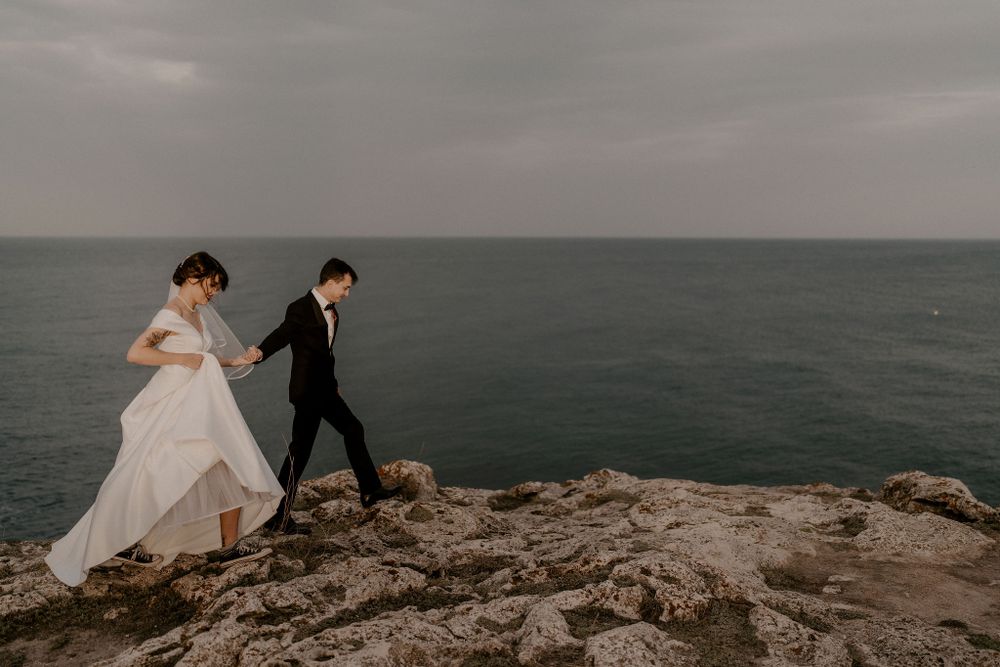 A romantic wedding couple stands on dramatic coastal cliffs overlooking the ocean at sunset.