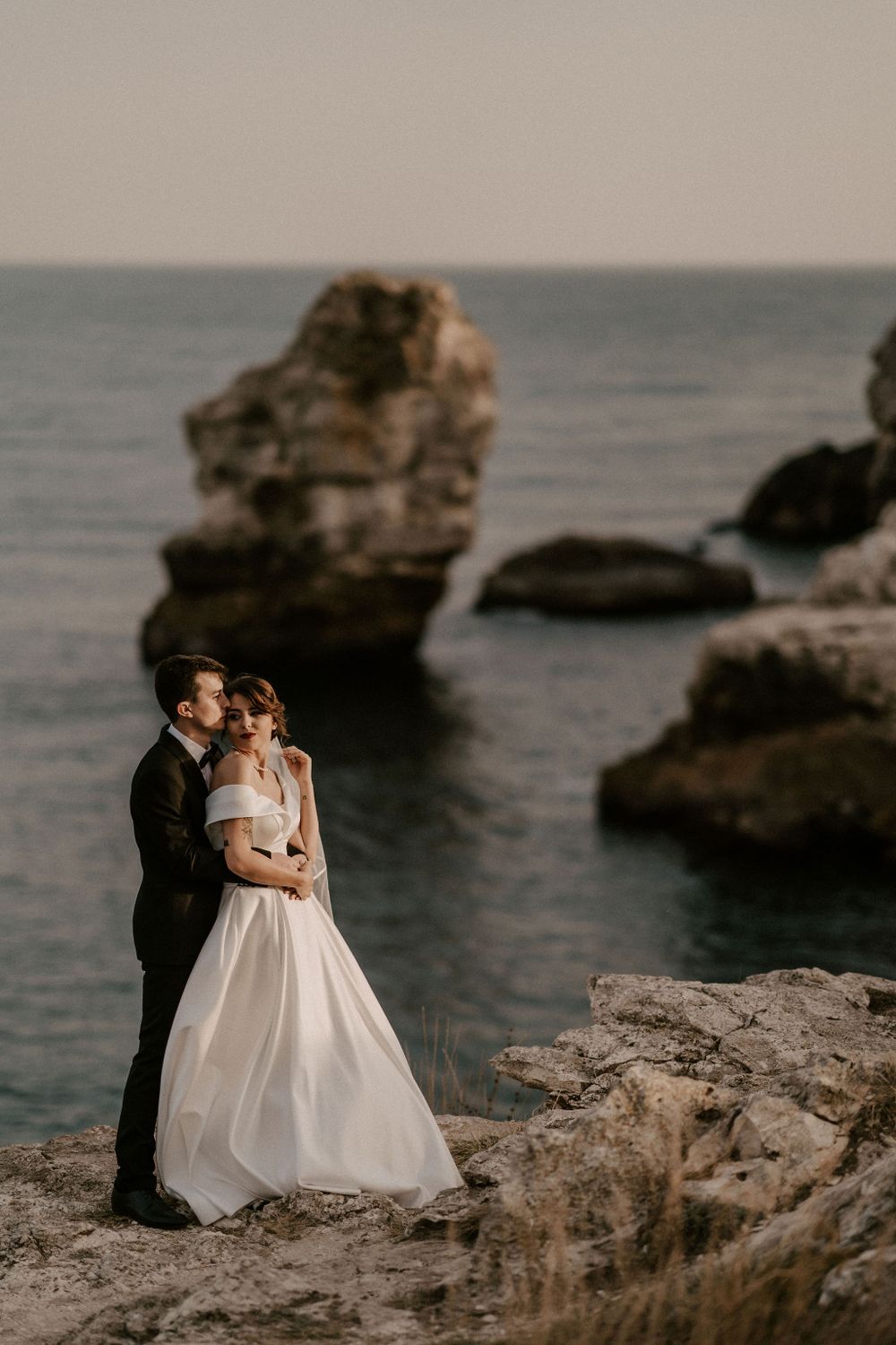 Newlyweds share an intimate moment on rugged ocean cliffs during golden hour.
