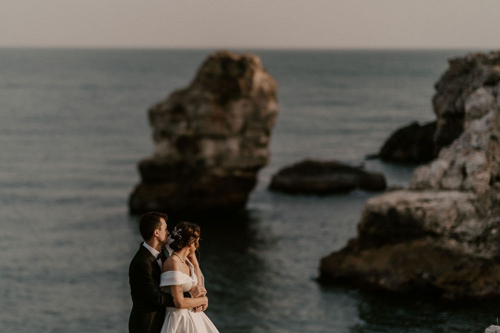 A wedding couple embraces on scenic coastal rocks with ocean waves in the background.