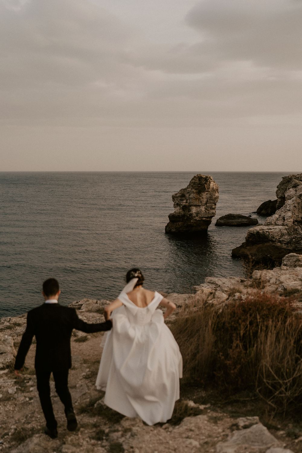 Newlyweds explore rocky coastline with dramatic ocean views and cloudy sky.
