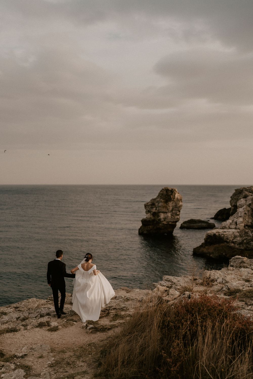 Wedding couple stands on cliff edge overlooking majestic ocean rock formations.