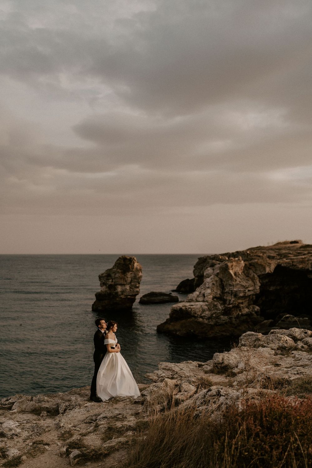 Romantic coastal wedding portrait with dramatic cloudy sky and rocky seascape.