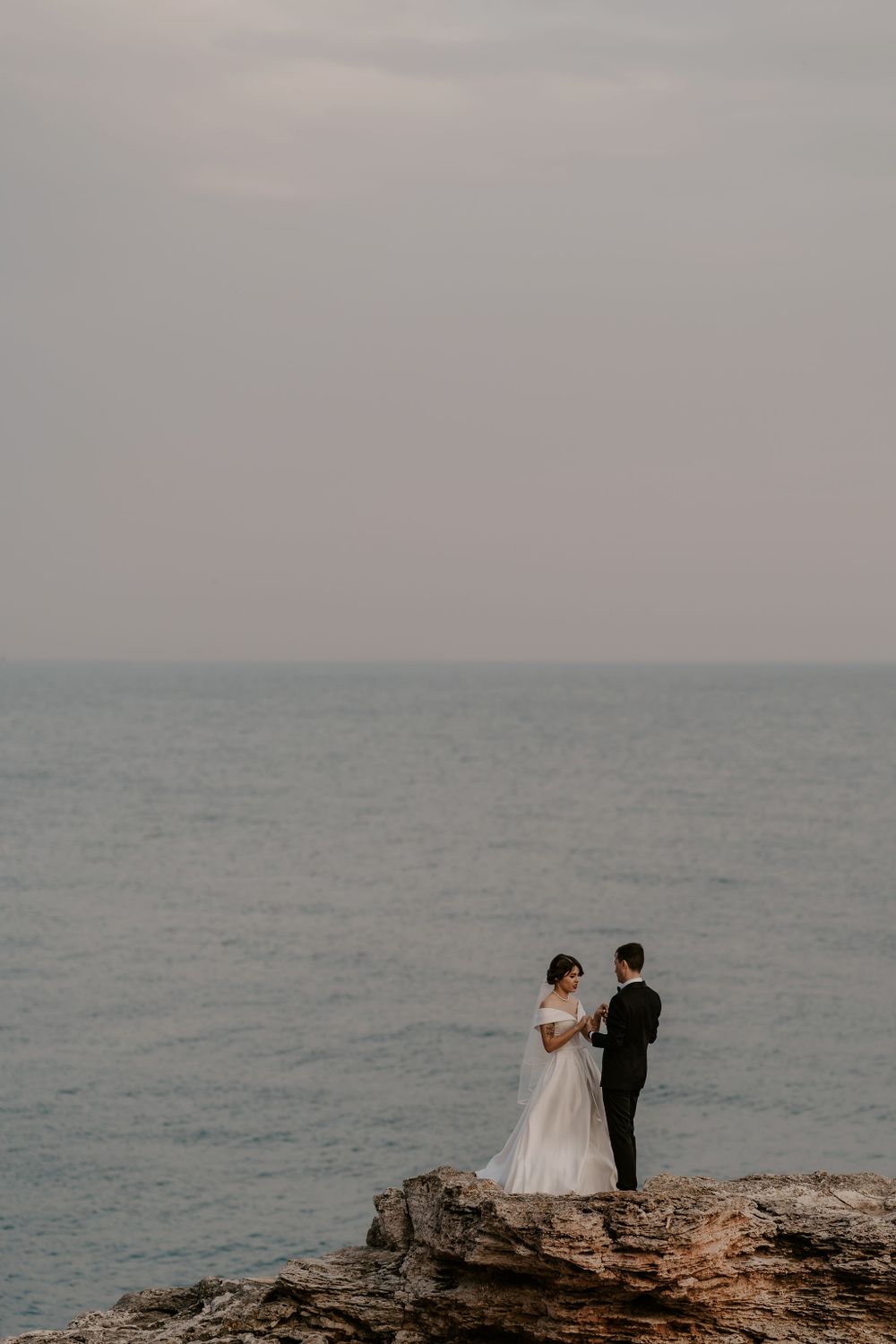 A couple in formal attire poses on rocky cliffs overlooking a serene ocean vista.