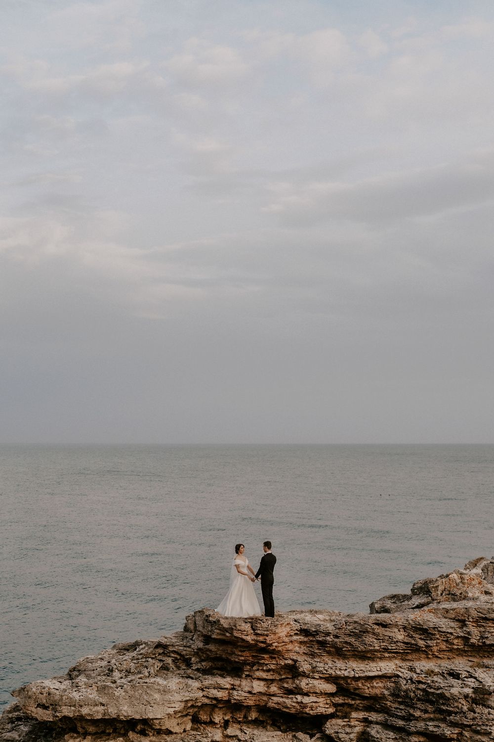 Two people in formal wear stand on dramatic ocean cliffs during an overcast day.