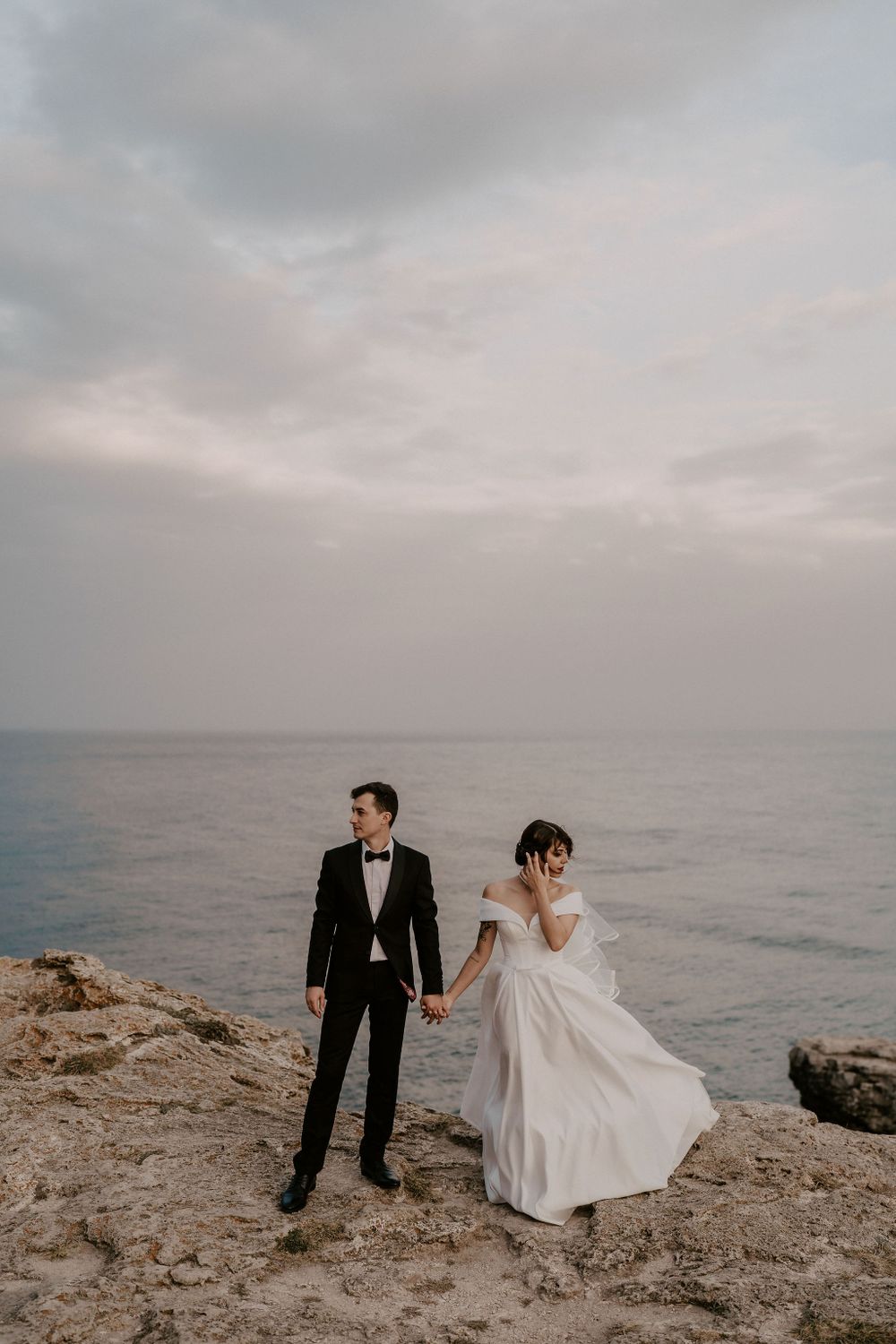 A couple in wedding attire explores rugged coastal cliffs by the ocean.
