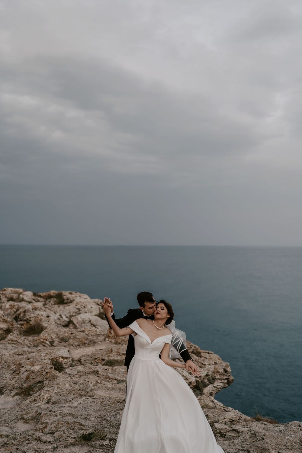 A couple in wedding attire poses dramatically on coastal cliffs overlooking the ocean.