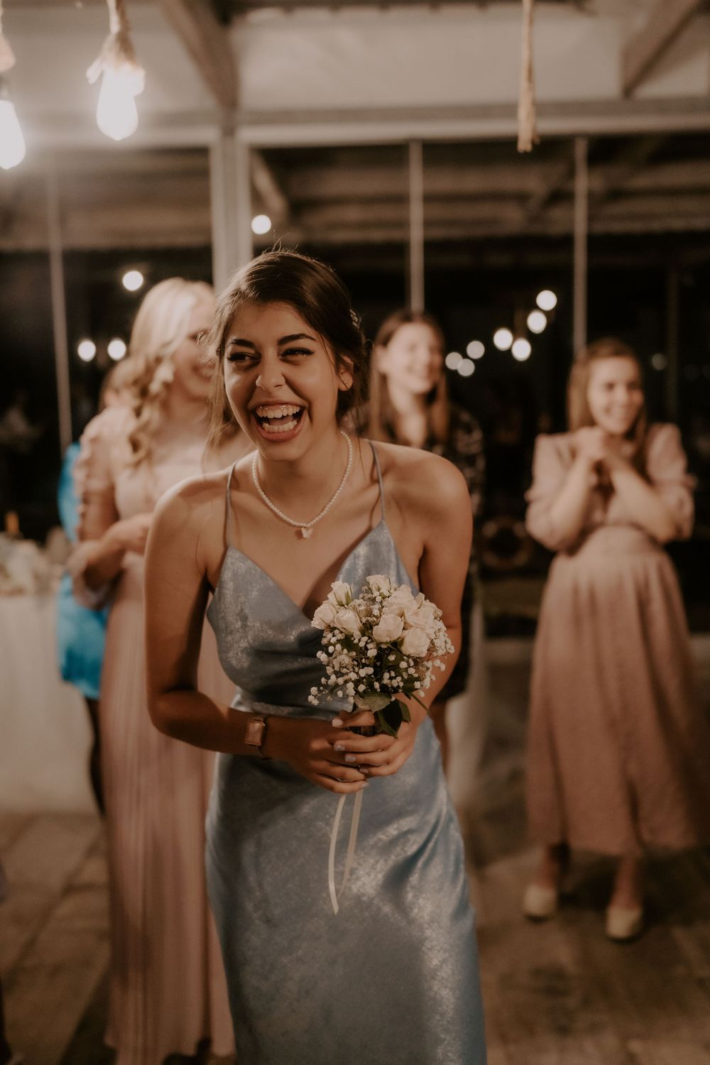 Bridesmaid in silver dress holds bouquet while laughing at wedding reception.