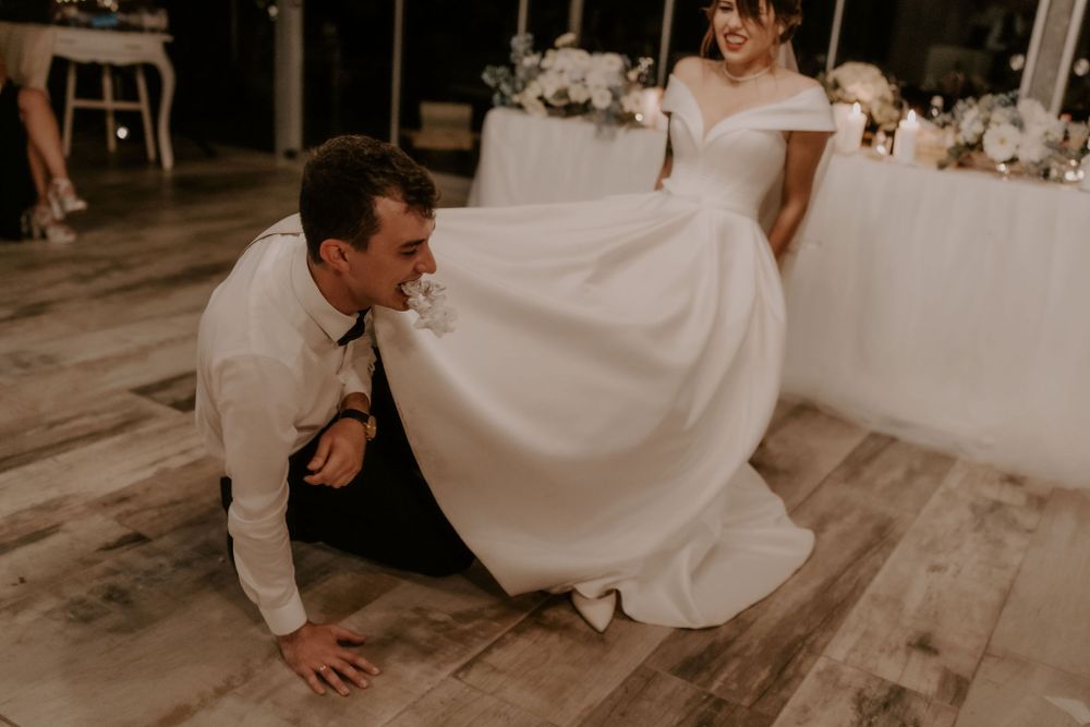 Couple shares playful moment during wedding dance on wooden floor with guests watching in background.