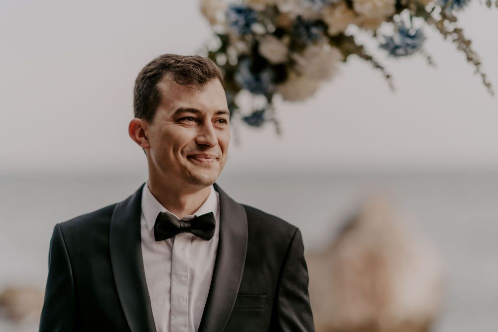 Groom in black tuxedo smiling at outdoor wedding venue.