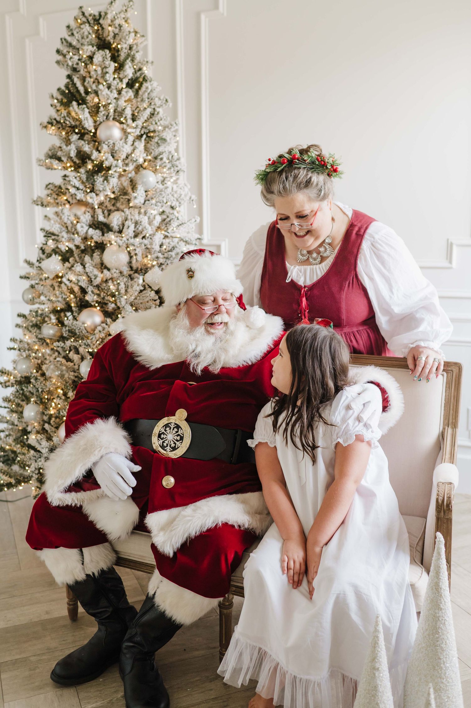 santa minis with santa and mrs claus with a little girl smiling behind a white christmas tree with natural light