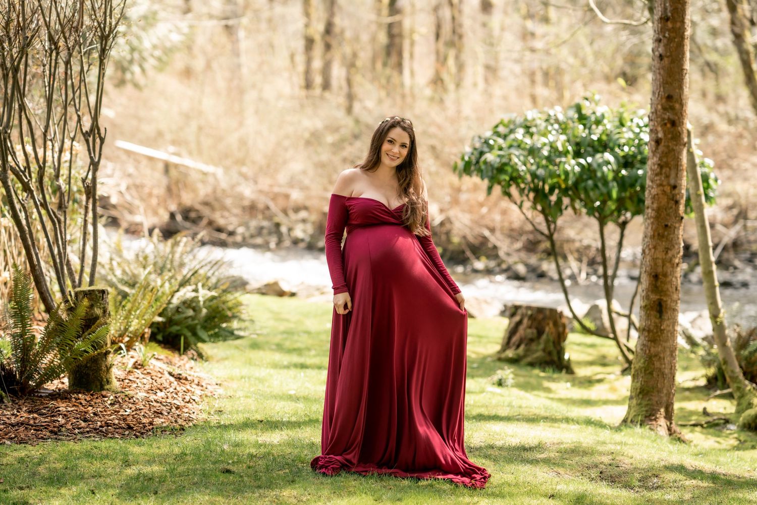 Expectant mother in burgundy dress poses in sunlit forest setting.