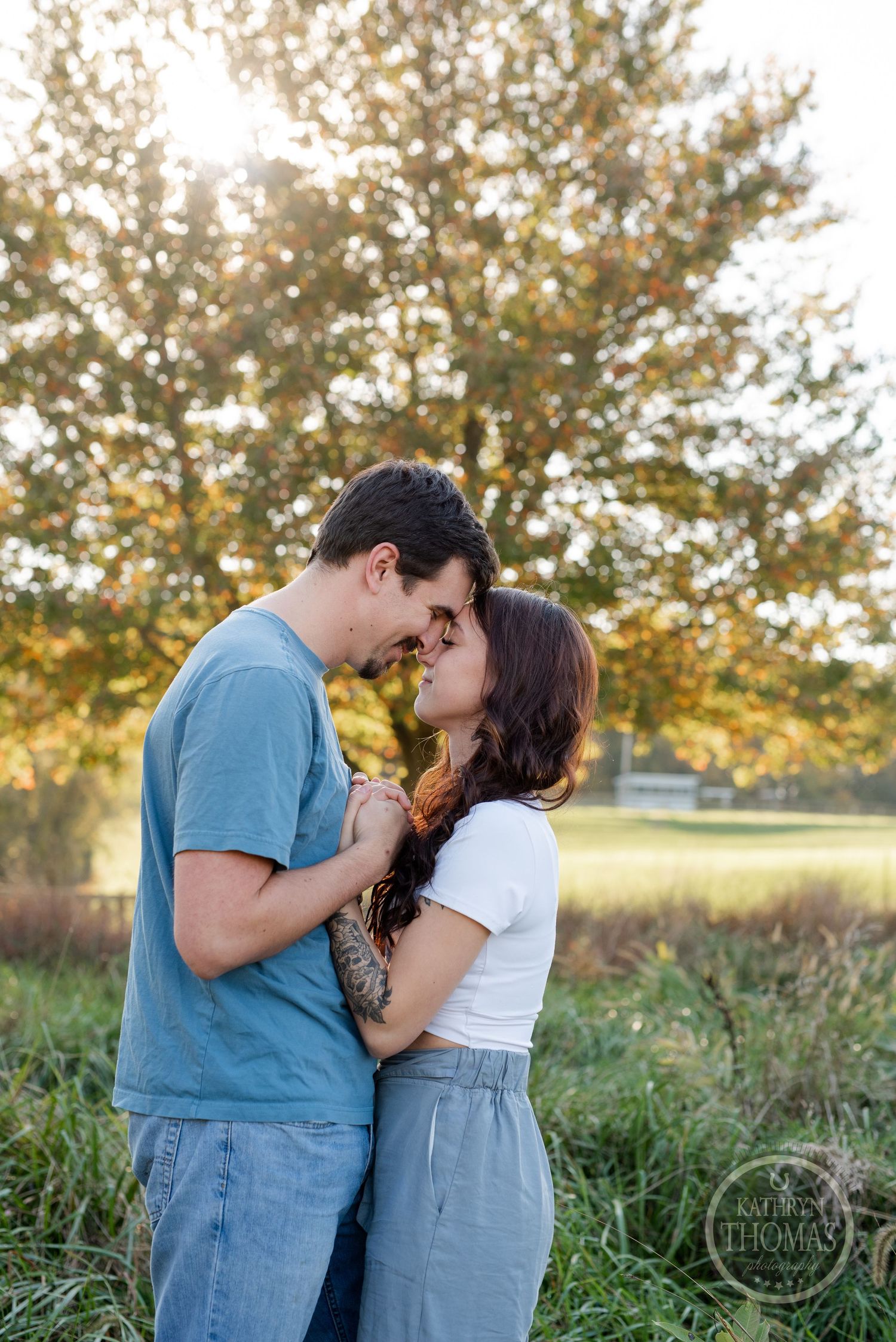 A tender embrace between two people standing in tall grass with autumn trees in the background.