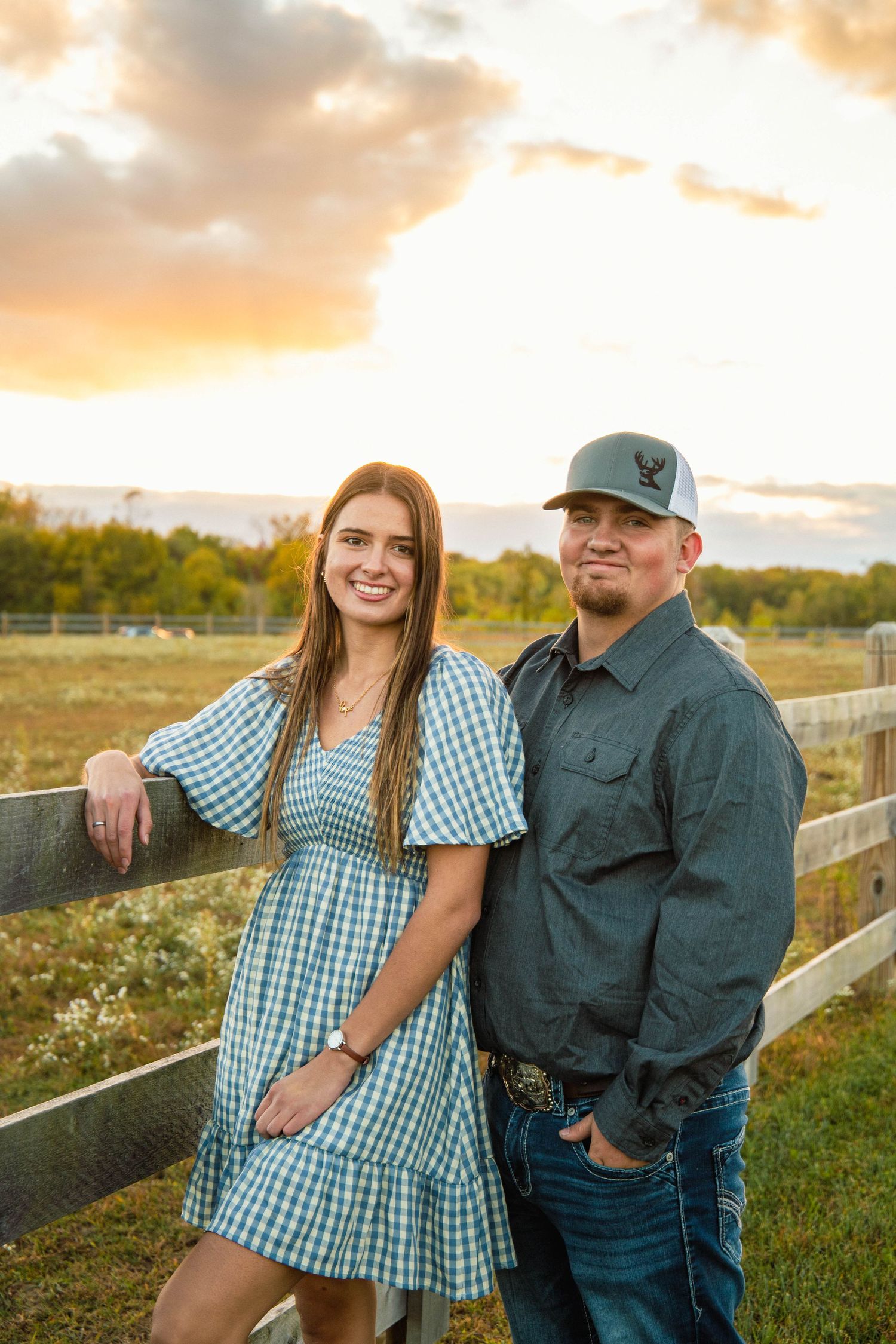 Couple posing together in a field at sunset with rustic wooden fence in background.