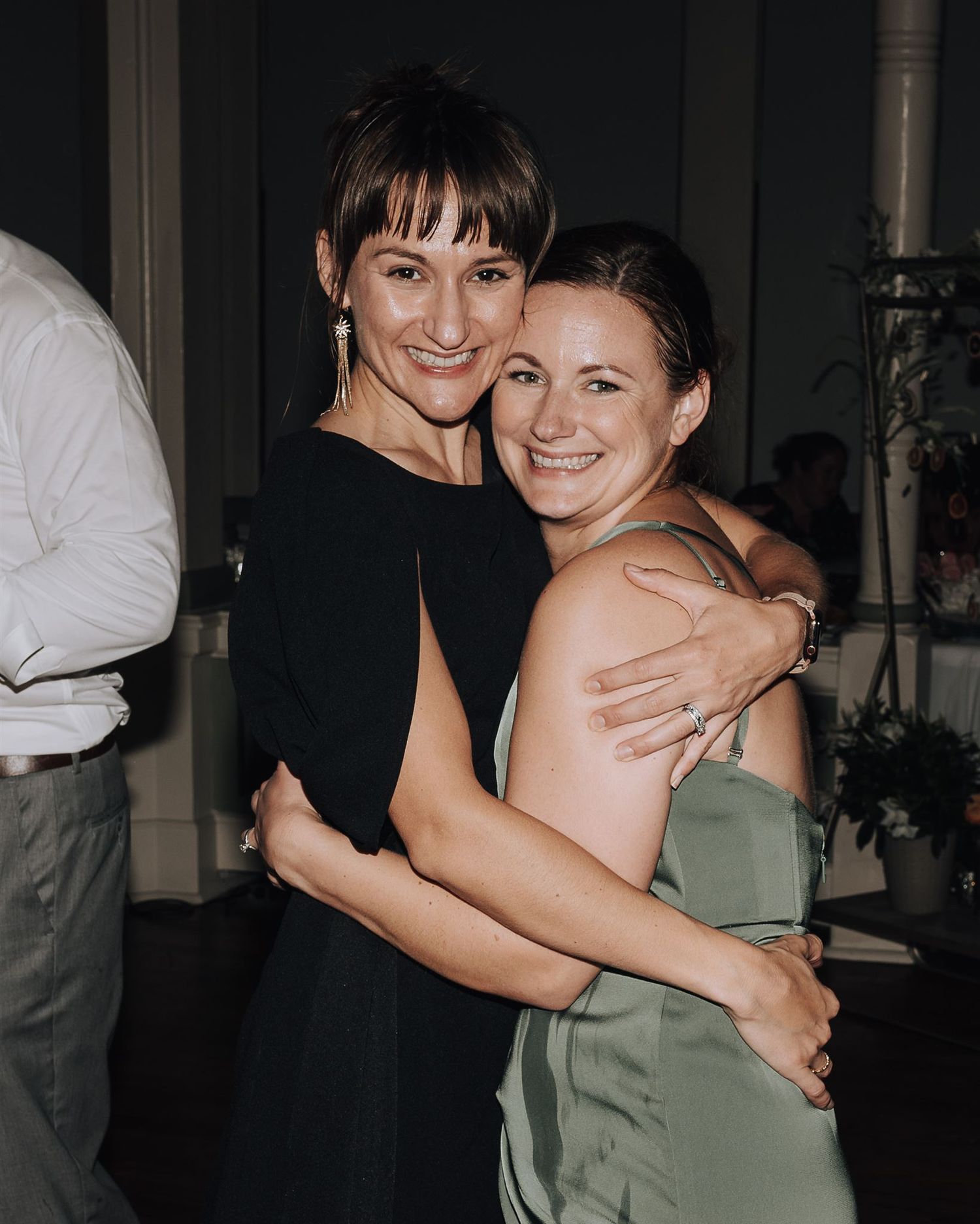 Two friends in black dresses sharing a warm embrace at a formal event.